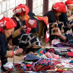 Women from the Red Dao tribe in traditional outfits working on cloth pieces for sale. The Red Dao are known for their intricate hand-sewn embroidery. You typically find them working and socializing in small groups on the sidewalks around town. Discovered by Mark Rentz at Cha Pai, Vietnam